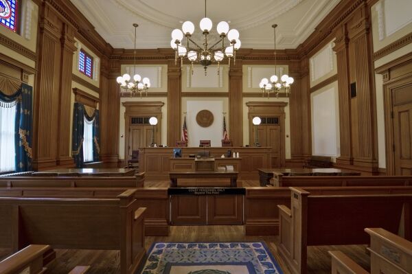 A wooden courtroom interior featuring a judge's bench, seating areas for the public, and decorative lighting with stained glass windows.