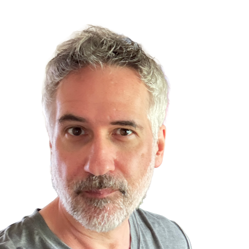 A man with gray hair and a beard, gazing directly at the camera in a well-decorated room featuring plants and musical instruments.