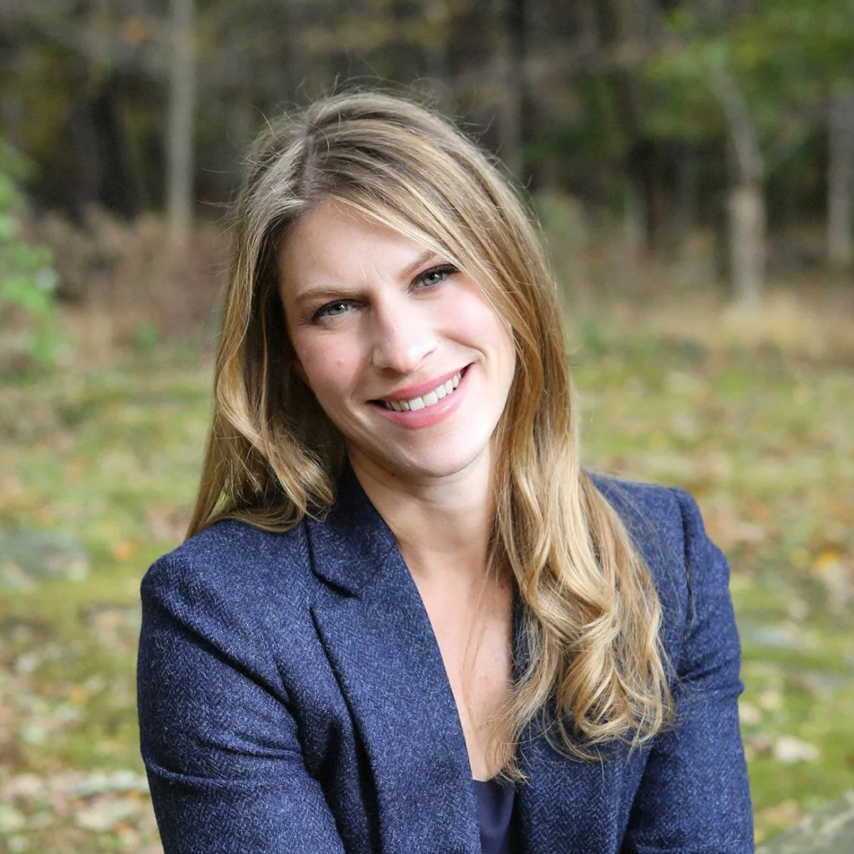A woman with long, wavy hair smiles warmly while seated in a forested area, wearing a dark blazer against a backdrop of soft focus greenery.